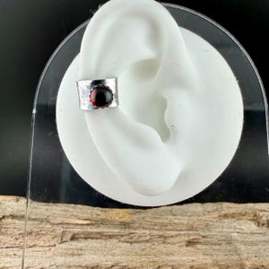 Puede incluir: Un brazalete de oreja plateado con textura martillada y una gema redonda de color rojo intenso. El brazalete est&aacute; dise&ntilde;ado para envolver el cart&iacute;lago de la oreja. La joya se exhibe en un modelo de oreja blanca.