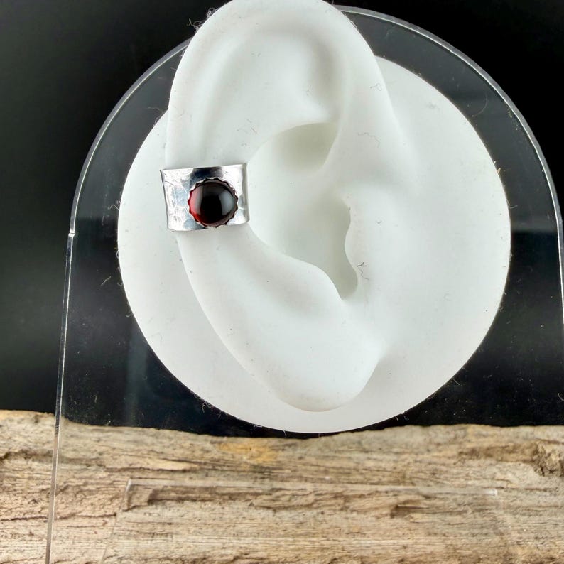 Puede incluir: Un brazalete de oreja plateado con textura martillada y una gema redonda de color rojo intenso. El brazalete est&aacute; dise&ntilde;ado para envolver el cart&iacute;lago de la oreja. La joya se exhibe en un modelo de oreja blanca.