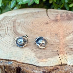 May include: A pair of silver-coloured cowboy hat stud earrings. The earrings feature a hat brim and a central indentation. The earrings are displayed on a wooden surface, with green foliage in the background.