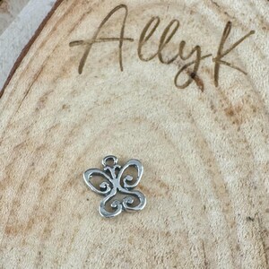 May include: A small, silver-colored butterfly charm with intricate openwork details. The charm is set against a light brown wooden surface with the handwritten text "Ally k" in the upper right corner.