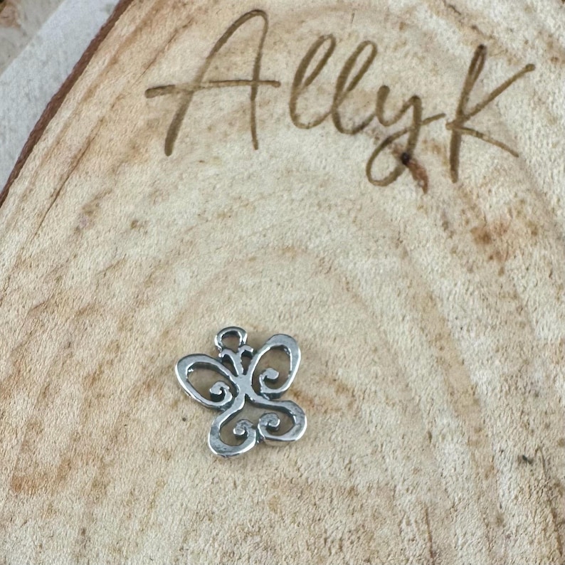 May include: A small, silver-colored butterfly charm with intricate openwork details. The charm is set against a light brown wooden surface with the handwritten text "Ally k" in the upper right corner.