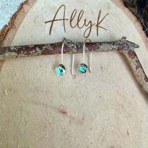 May include: A pair of silver drop earrings featuring small, round, turquoise stones with brown matrix veining. The earrings have a simple hook design and are displayed hanging from a thin, dark brown twig against a light wood background.