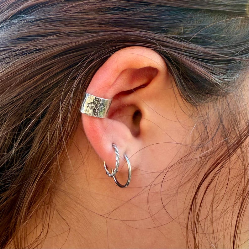Puede incluir: Un pendiente tipo ear cuff de plata con un patr&oacute;n de cruz geom&eacute;trica estampado en el cart&iacute;lago superior, acompa&ntilde;ado de dos aros de plata en el l&oacute;bulo, uno con textura de cuerda trenzada y el otro con un acabado liso y pulido.