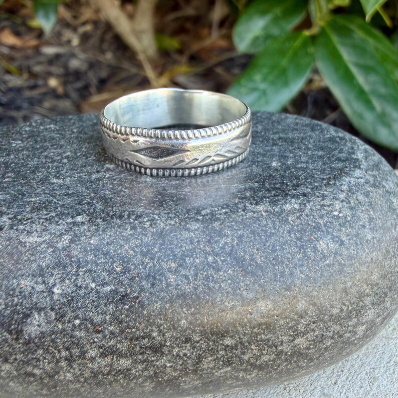 May include: A silver-toned metal ring featuring a band with a central row of etched diamond-shaped patterns and horizontal lines, bordered by two thin, raised, beaded edges that run along the top and bottom circumference of the band.
