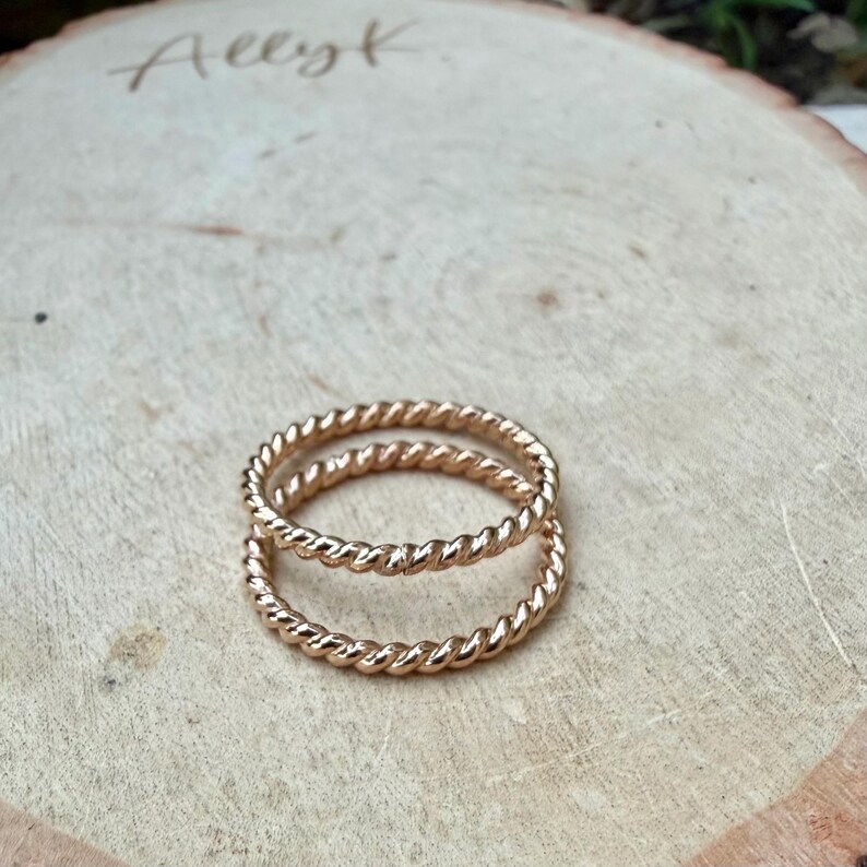 May include: Two gold-colored, twisted rope rings are stacked on a light brown wooden surface. The rings have a textured, braided appearance. The rings are the focal point, with the word "Allyk" handwritten in cursive in the upper left corner.