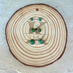 May include: Three pairs of round stud earrings featuring polished green turquoise stones set in silver metal. The earrings are arranged in a vertical column on a circular wooden slice with a laser-engraved logo in the center.