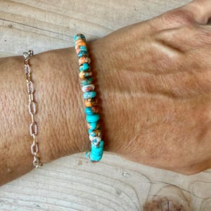 May include: A beaded bracelet featuring small, round, disc-shaped beads in vibrant turquoise, white, and orange hues, worn alongside a delicate silver-toned chain bracelet with oval-shaped links. The beads display natural color variations.