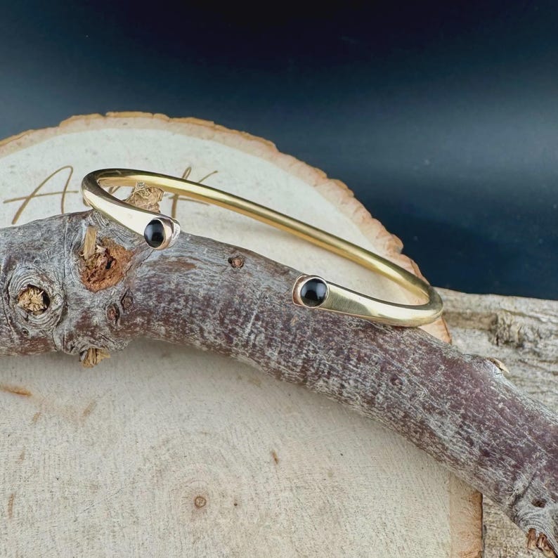 May include: A gold-colored bangle bracelet with two small, round, black stones set at the ends. The bracelet is displayed on a piece of wood with a natural, light brown color, against a dark background.
