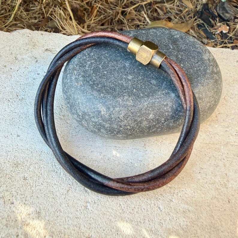 May include: A multi-strand bracelet featuring two dark brown leather cords twisted together. The bracelet is secured with a cylindrical brass magnetic clasp that has a smooth, metallic finish and a distinct hexagonal shape on the side.