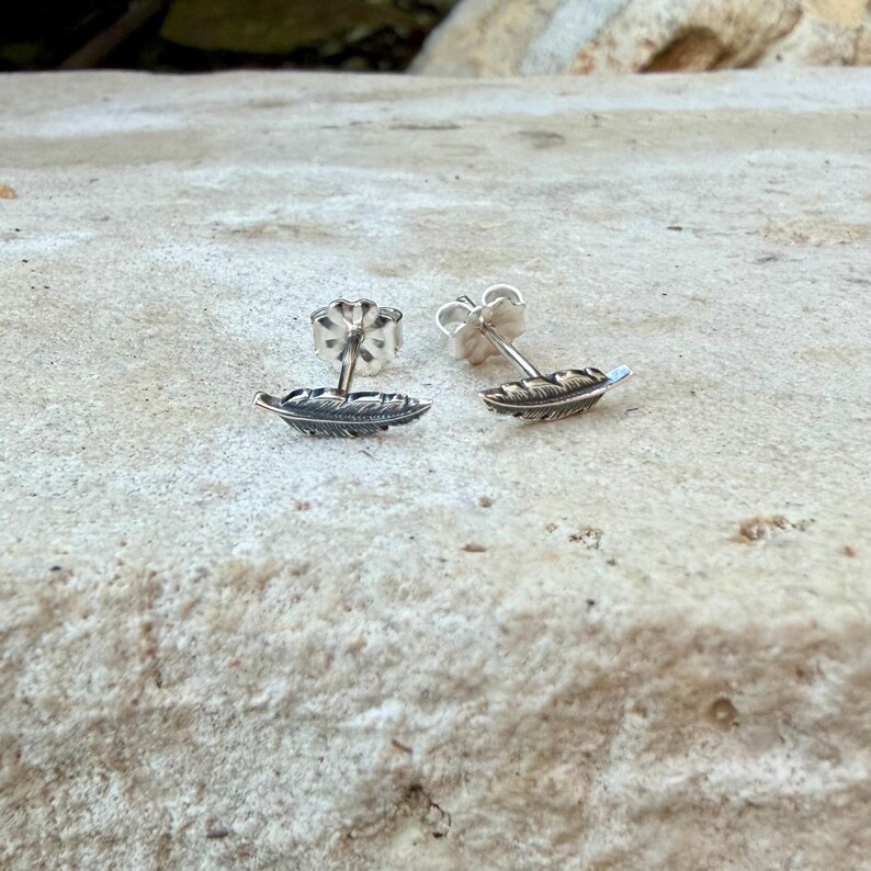May include: A pair of silver feather stud earrings. Each earring features a detailed feather design with a post and butterfly backing. The earrings are displayed on a textured, light-colored surface.