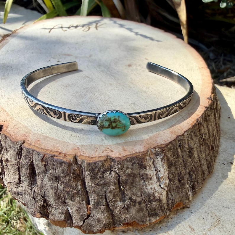 Op de afbeelding: Een zilveren manchetarmband met een ovale turkoois steen. De armband heeft een ge&euml;tst ontwerp langs de band. De turkoois steen heeft een levendige blauwgroene kleur. De armband wordt gepresenteerd op een houten oppervlak.
