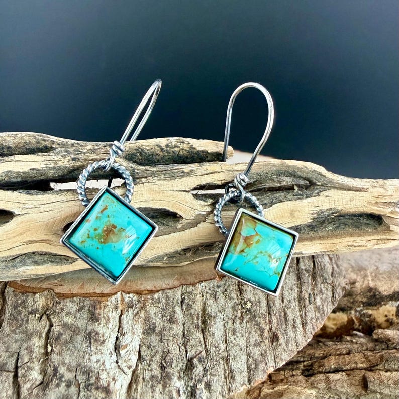 May include: A pair of silver dangle earrings with square turquoise stones. The turquoise stones are set in a silver frame and have brown and tan markings. The earrings are displayed on a piece of driftwood.