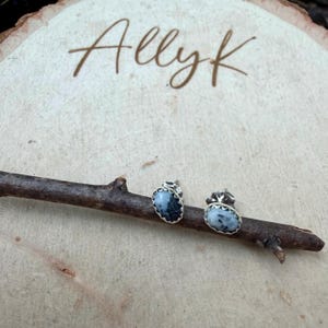 May include: A pair of silver stud earrings with oval, black and white speckled stones. The earrings are set on a wooden twig, with the word "Allyk" written in cursive on a wooden background.