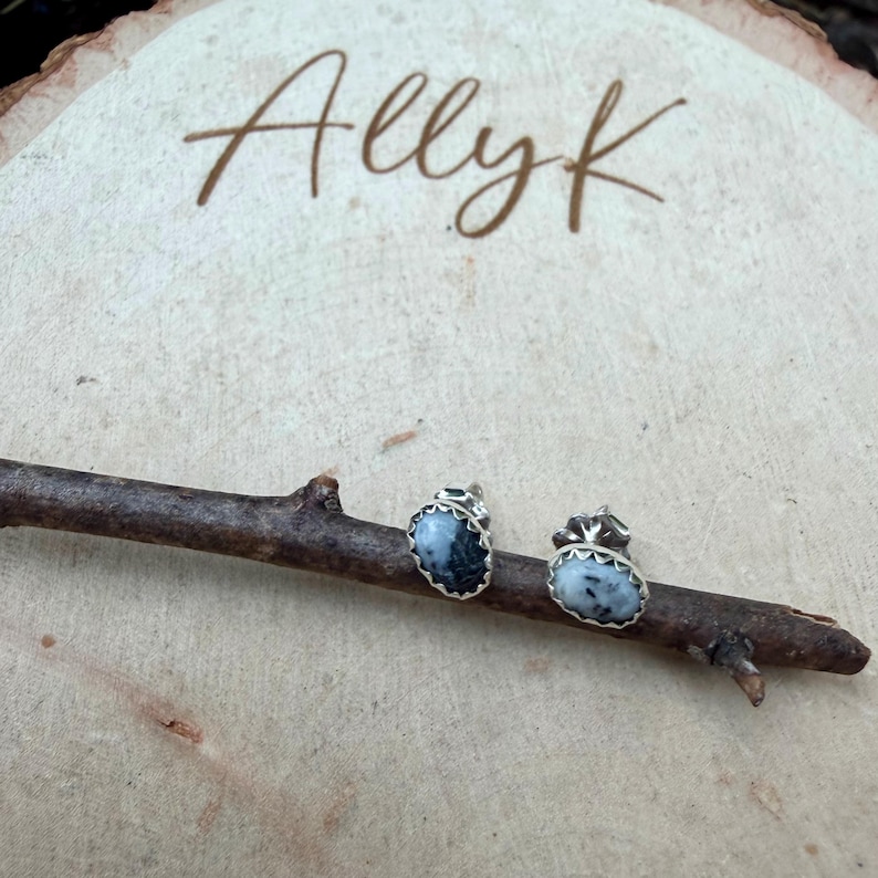 May include: A pair of silver stud earrings with oval, black and white speckled stones. The earrings are set on a wooden twig, with the word "Allyk" written in cursive on a wooden background.