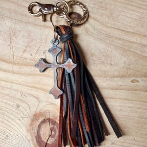 Op de afbeelding: Een sleutelhanger met een metalen kruisbedel met een verweerde bruine en zilveren afwerking, bevestigd aan een bundel dunne, gelaagde leren stroken in zwart- en bruintinten. Het geheel is voorzien van een zilverkleurige karabijnhaak en een gedraaide ring.