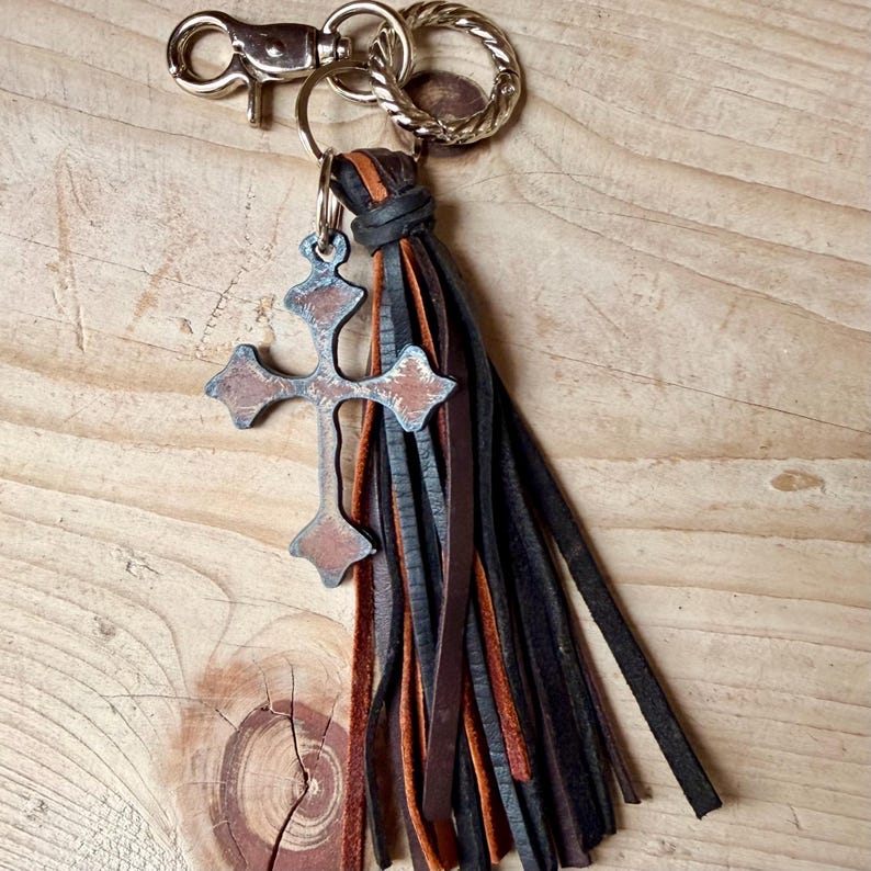 Op de afbeelding: Een sleutelhanger met een metalen kruisbedel met een verweerde bruine en zilveren afwerking, bevestigd aan een bundel dunne, gelaagde leren stroken in zwart- en bruintinten. Het geheel is voorzien van een zilverkleurige karabijnhaak en een gedraaide ring.