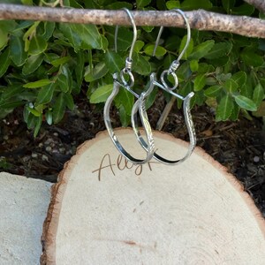 May include: A pair of silver hoop earrings with a textured design, hanging from a branch against a backdrop of green foliage. The earrings are displayed on a wooden slice with the handwritten word "Ally".