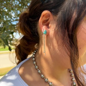 May include: A silver earring featuring a teardrop-shaped turquoise stone with a dark matrix, accented by two delicate silver chains hanging from the base. The earring is worn alongside a small silver hoop and a necklace made of large silver beads.
