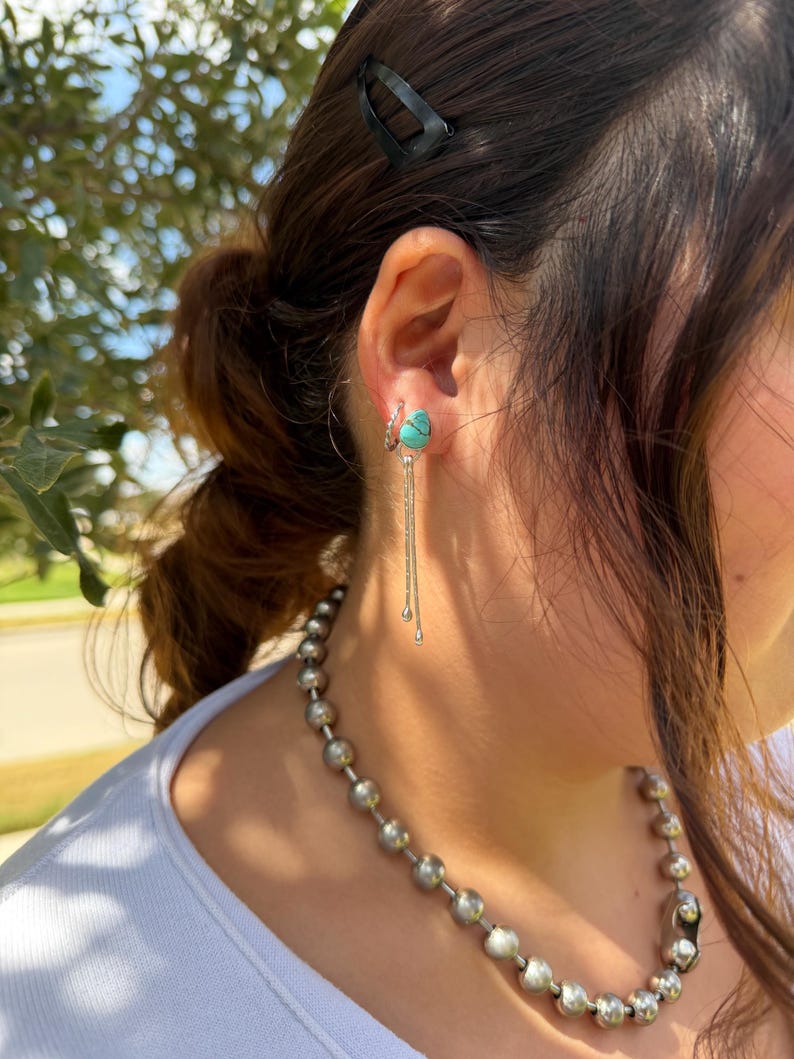 May include: A silver earring featuring a teardrop-shaped turquoise stone with a dark matrix, accented by two delicate silver chains hanging from the base. The earring is worn alongside a small silver hoop and a necklace made of large silver beads.