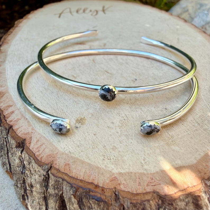May include: Two silver-toned cuff bracelets with small, round, black and white speckled stones. The bracelets are open-ended and rest on a wooden surface. The word "Allyk" is handwritten on the wood.