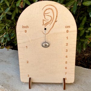 May include: A silver stud earring with a circular, sunburst-patterned face, displayed on a light-colored wooden stand featuring an engraved ear outline and a ruler with markings in inches and centimeters for scale and size reference.