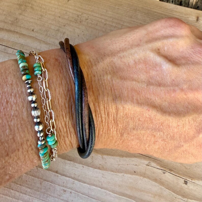 May include: A layered bracelet set consisting of a dark brown leather cord wrap bracelet and a multi-strand bracelet featuring turquoise beads, small silver-toned metal beads, and a delicate silver-toned chain with elongated oval links.