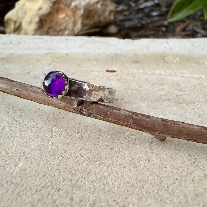 May include: A silver ring with a textured band and a large, round, deep purple amethyst gemstone. The ring is set on a natural wooden twig against a neutral background. The gemstone is the focal point.