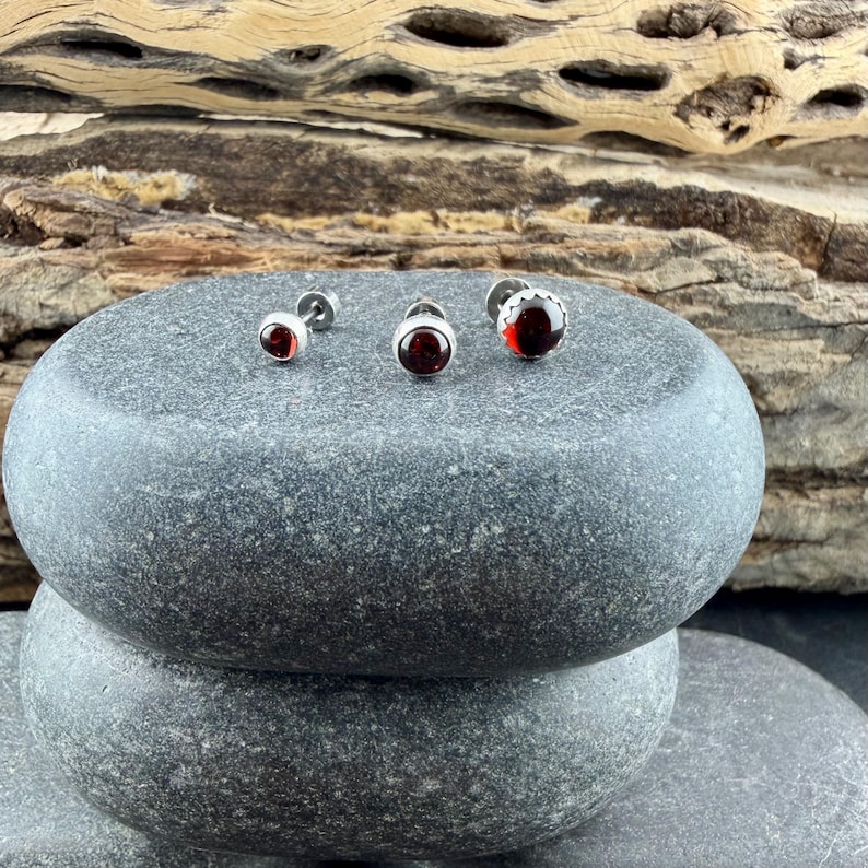May include: Three stud earrings featuring round, deep red garnet stones set in polished silver-toned metal settings. The earrings are arranged in a horizontal line, showcasing the dark red gemstone cabochons and the metallic circular frames.