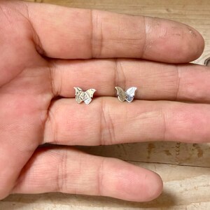 K&ouml;nnte beinhalten: Ein Paar kleine, silberfarbene Schmetterlings-Ohrstecker. Jeder Ohrstecker weist ein detailliertes, texturiertes Fl&uuml;gelmuster mit einer polierten metallischen Oberfl&auml;che auf und ist als passendes Set konzipiert.