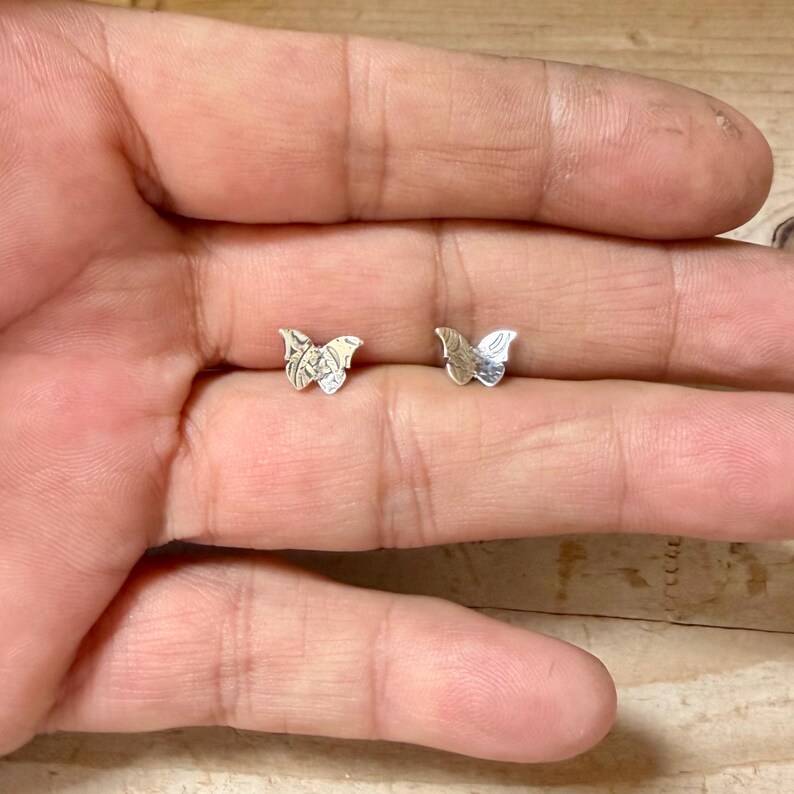K&ouml;nnte beinhalten: Ein Paar kleine, silberfarbene Schmetterlings-Ohrstecker. Jeder Ohrstecker weist ein detailliertes, texturiertes Fl&uuml;gelmuster mit einer polierten metallischen Oberfl&auml;che auf und ist als passendes Set konzipiert.