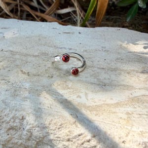 以下が含まれることがあります： ハンマー仕上げの金属テクスチャが施されたシルバーのオープンリング。バンドの両端には小さな丸い深紅のガーネットの宝石がセットされており、石に光沢のある仕上げが施されたバイパススタイルのデザインになっています。