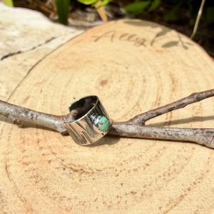 May include: A silver-toned ring with a turquoise stone set in a wide band. The ring has a textured surface and an open design. The ring is displayed on a small branch against a wooden background.