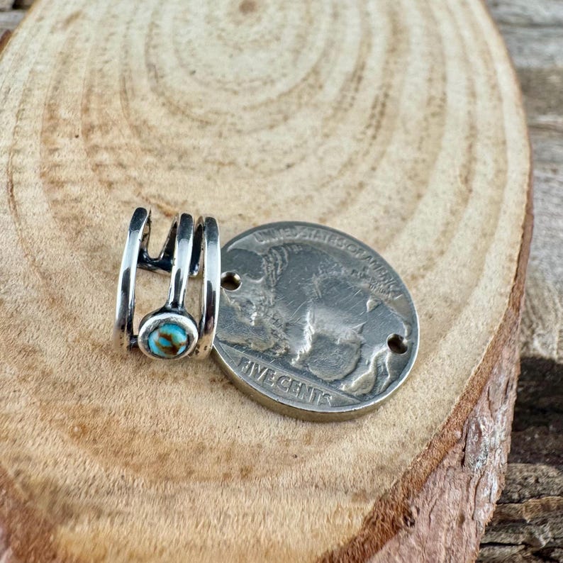 May include: A silver-toned ring with a small, round turquoise stone. The ring is next to a silver coin with a buffalo image. The ring has a unique, open design. The coin reads "FIVE CENTS".