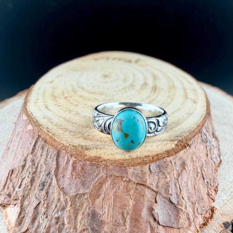 May include: A silver ring with an oval turquoise stone. The ring has a detailed floral design on the band. The turquoise stone is a vibrant blue-green color with brown matrix patterns. The ring is displayed on a wooden surface.