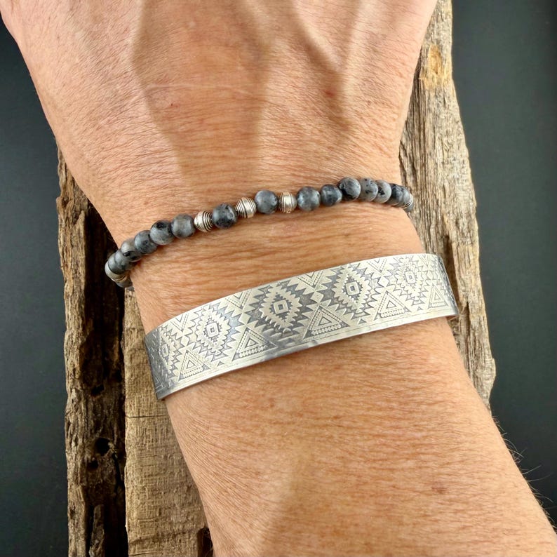 May include: A silver cuff bracelet featuring an engraved geometric pattern of triangles and diamonds, paired with a beaded bracelet made of small grey stone spheres and interspersed with polished silver-toned metal spacer beads.
