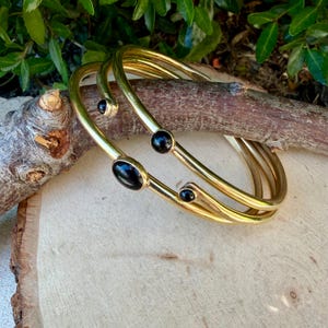 May include: Three gold-colored bangles with black oval and round stones. The bangles are stacked on top of each other, creating a layered effect. The jewelry is displayed on a wooden surface.