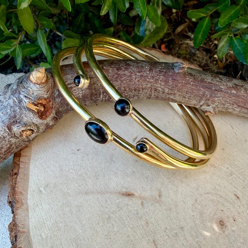 May include: Three gold-colored bangles with black oval and round stones. The bangles are stacked on top of each other, creating a layered effect. The jewelry is displayed on a wooden surface.