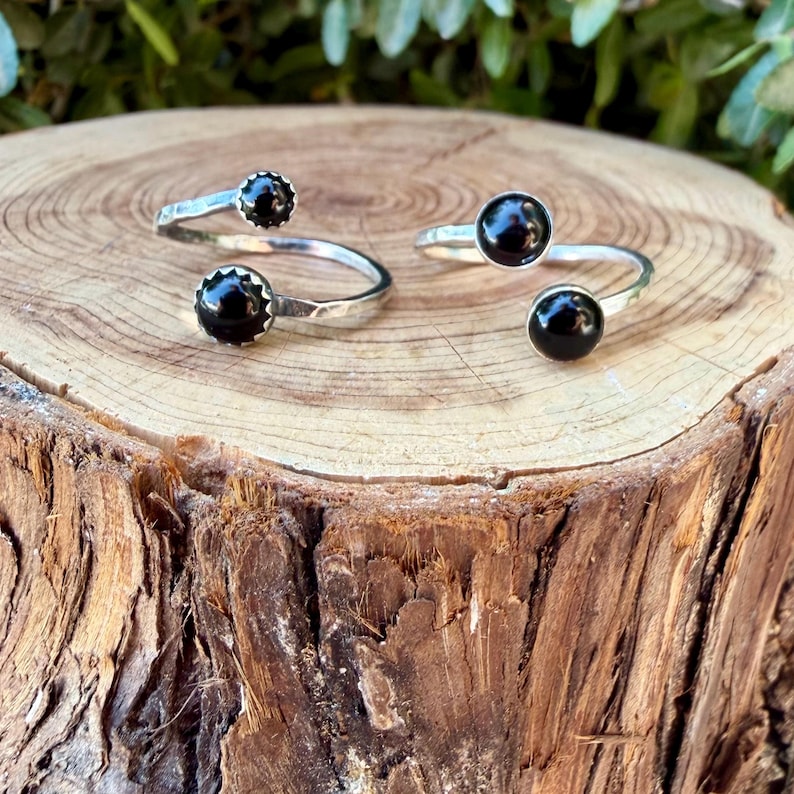 May include: Two open-style rings crafted from silver-toned metal, each featuring two round, polished black stones set at the ends of the band. The rings have a smooth, reflective finish and a simple, adjustable design with circular black accents.