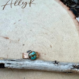 May include: A copper-colored ring with a large, round turquoise stone. The ring band has a textured, etched design. The turquoise stone is a mix of blue and brown. The ring is displayed on a piece of wood.