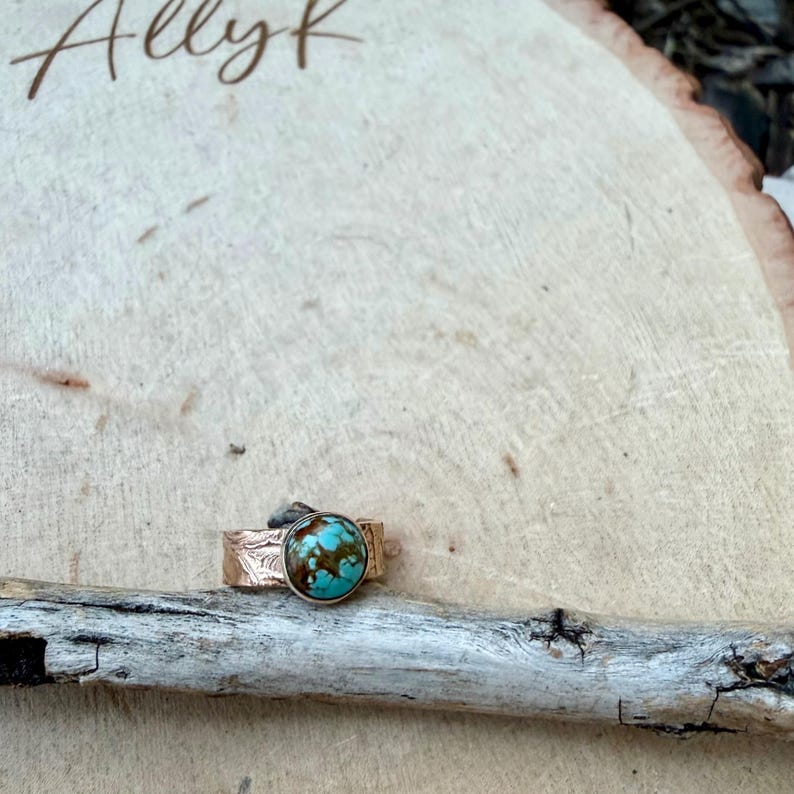 May include: A copper-colored ring with a large, round turquoise stone. The ring band has a textured, etched design. The turquoise stone is a mix of blue and brown. The ring is displayed on a piece of wood.