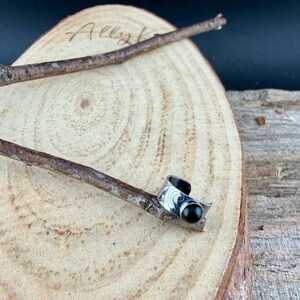 May include: A silver-toned ring with a black gemstone, resting on a wooden surface. The ring features an engraved design and a curved shape. The word "Ally" is visible on the wooden surface.