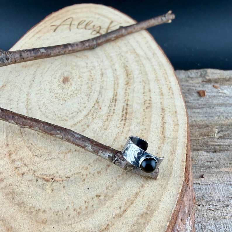 May include: A silver-toned ring with a black gemstone, resting on a wooden surface. The ring features an engraved design and a curved shape. The word "Ally" is visible on the wooden surface.