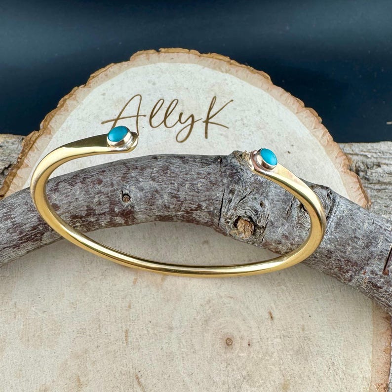 May include: A gold-colored cuff bracelet with turquoise-colored stones at each end. The bracelet is curved and open, with the text "Allyk" in cursive on a wooden background.