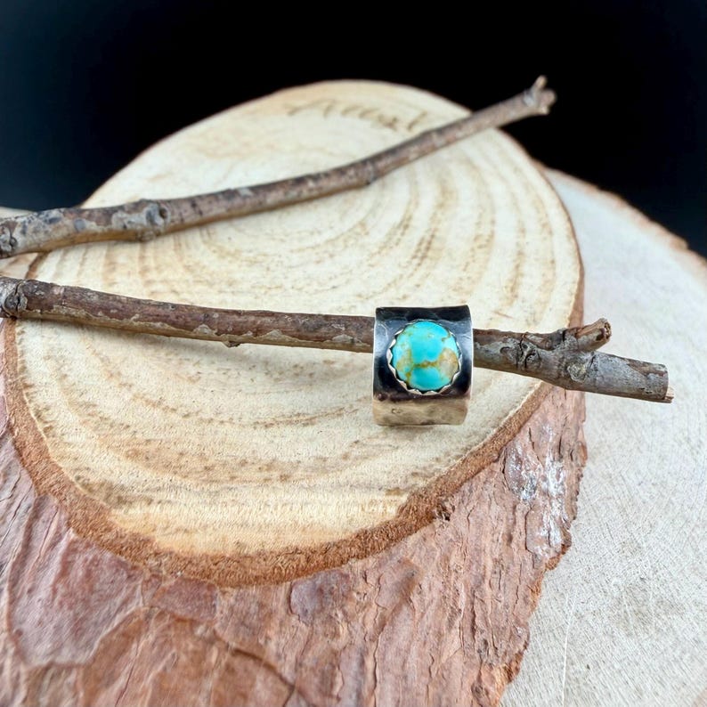 May include: A silver ring with a square band and a round, turquoise stone. The ring is displayed on a small branch, set against a wooden background. The turquoise stone has brown and gold accents.