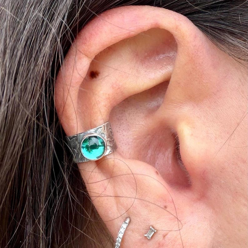 May include: A silver ear cuff with an embossed floral design and a round, emerald-green gemstone. The cuff is worn on the ear, showcasing the intricate details and vibrant color of the stone.
