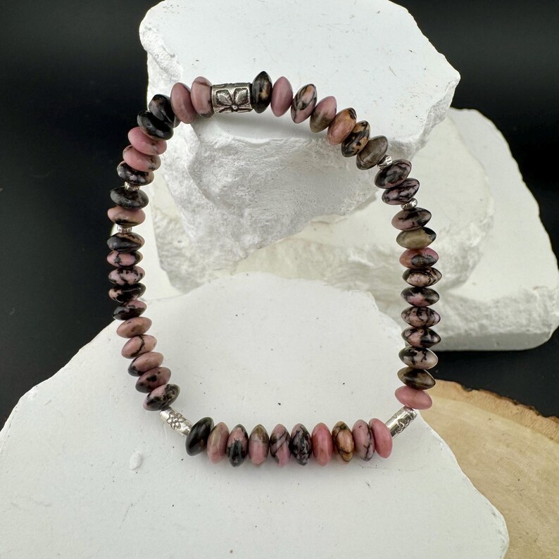 May include: A beaded bracelet featuring a mix of pink, brown, and black stones. The bracelet is strung on a silver-colored wire and has a silver clasp. The stones are oval-shaped and vary in size. The bracelet is displayed on a white surface.