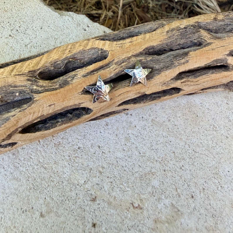 May include: Two small, five-pointed star-shaped stud earrings with a polished, reflective silver-tone metallic finish. Each star features a subtle, textured surface pattern that catches the light, creating a bright, gleaming appearance.
