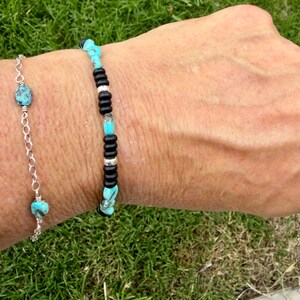 May include: Two bracelets are shown. One is a silver chain with turquoise beads. The other bracelet has black and silver beads with turquoise accents. The bracelets are on a wrist, with green grass in the background.