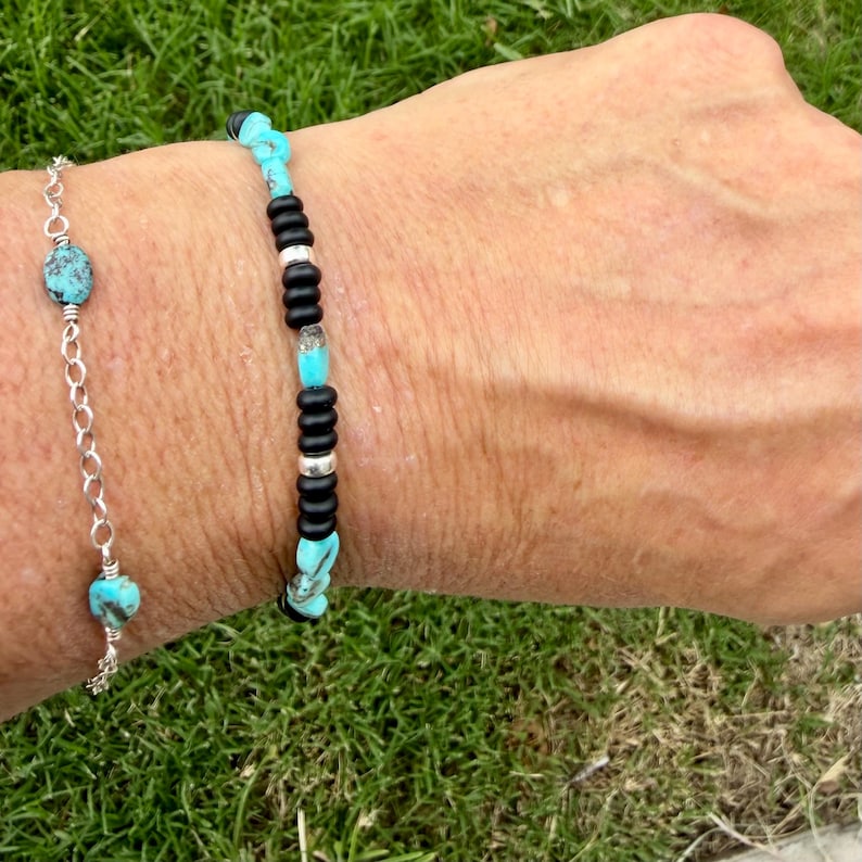 May include: Two bracelets are shown. One is a silver chain with turquoise beads. The other bracelet has black and silver beads with turquoise accents. The bracelets are on a wrist, with green grass in the background.