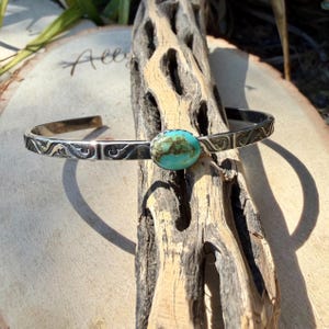 Op de afbeelding: Een zilveren manchetarmband met een ovale turkoois steen. De armband heeft een gegraveerd patroon langs de randen. De turkoois steen heeft een levendige blauwgroene kleur met bruine marmering. De armband wordt getoond op een stuk hout.